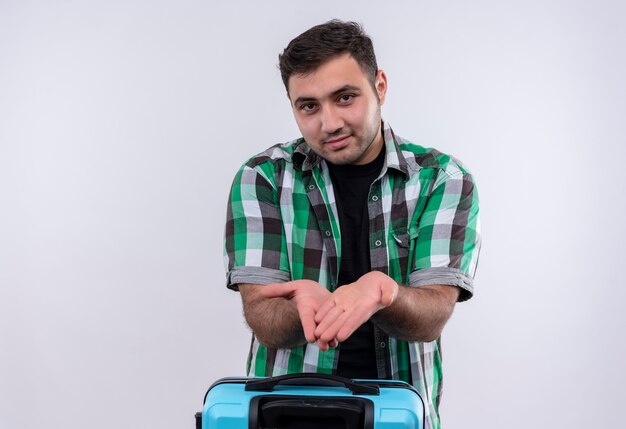 Young traveler man in checked shirt holding suitcase showing hands asking for money standing over white wall