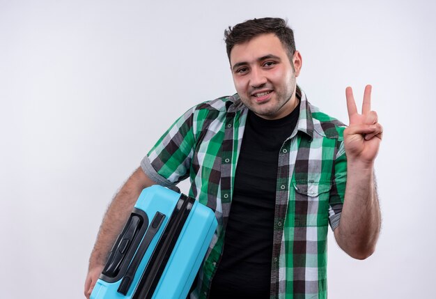 Young traveler man in checked shirt holding suitcase smiling and showing number three with fingers standing over white wall