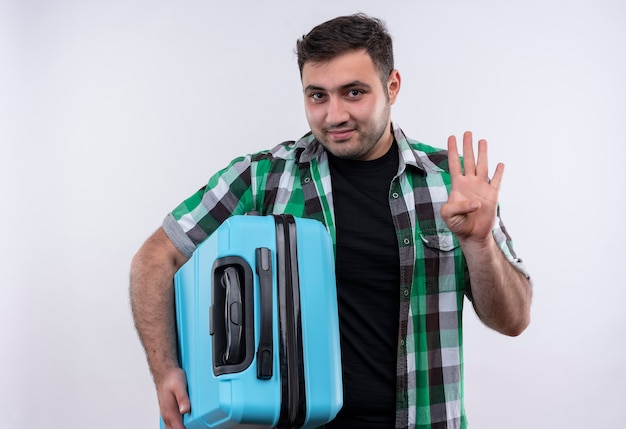 Free photo young traveler man in checked shirt holding suitcase smiling showing and pointing up with fingers number four standing over white wall