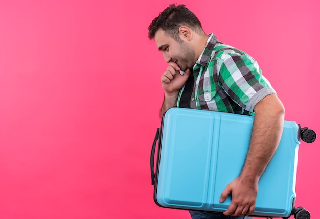 ピンクの壁の上の顔に物思いにふける表情で横に立っているスーツケースを保持しているチェックシャツの若い旅行者の男