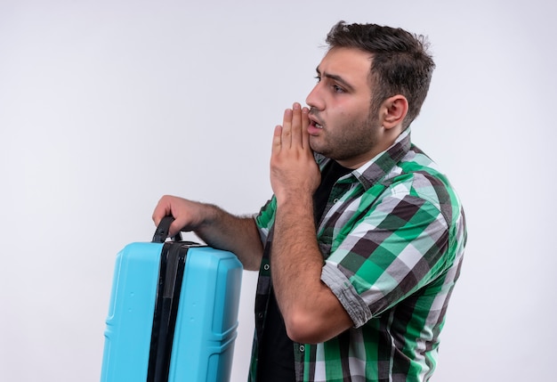 Young traveler man in checked shirt holding suitcase telling a secret with hand near his mouth standing sideways white wall