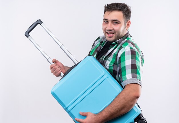 Young traveler man in checked shirt holding suitcase using as a guitar feeling joyfull, having fun standing over white wall