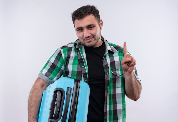 Молодой путешественник в клетчатой рубашке держит чемодан с уверенной улыбкой на лице, показывая указательный палец, стоящий над белой стеной