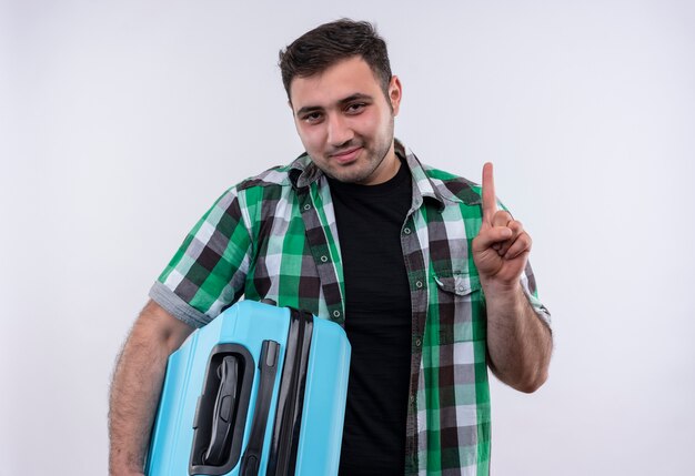 Young traveler man in checked shirt holding suitcase with confident smile on face showing index finger standing over white wall