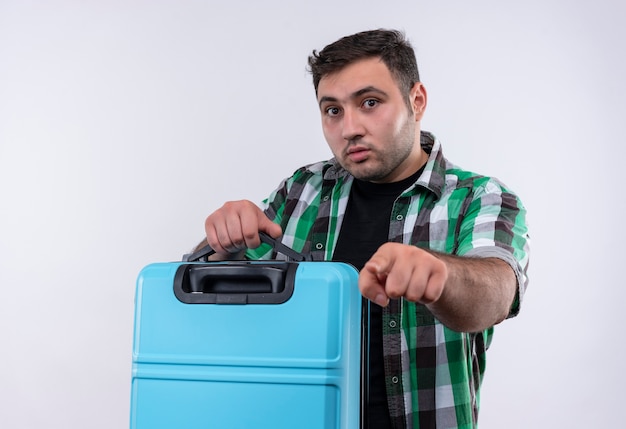 Uomo giovane viaggiatore in camicia a quadri tenendo la valigia con espressione di paura che punta con il dito indice, in piedi sopra il muro bianco