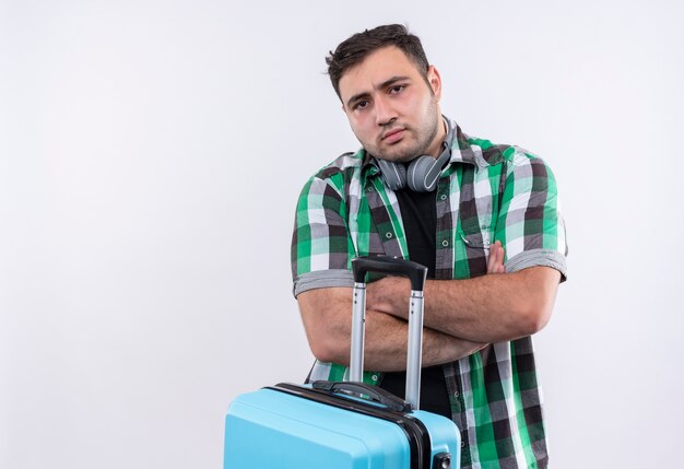 Young traveler man in checked shirt holding suitcase with frowning face standing over white wall