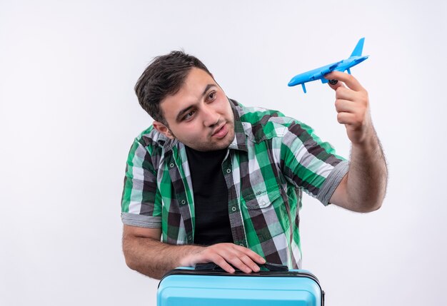 Young traveler man in checked shirt standing with suitcase holding toy airplane smiling with happy face over white wall