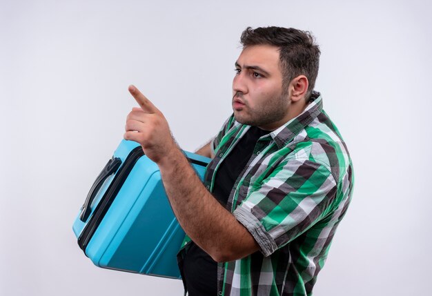 Молодой путешественник в клетчатой рубашке стоит с чемоданом, указывая пальцем в сторону, выглядит смущенным над белой стеной