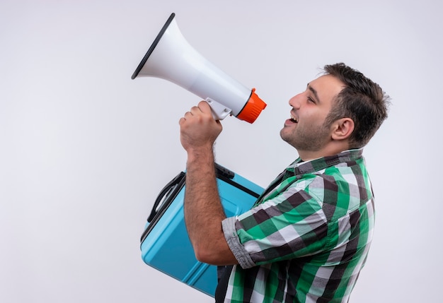 Young traveler man in checked shirt standing with suitcase shouting to megaphone over white wall