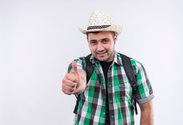 Young traveler man in checked shirt and summer hat with backpack smiling with happy face showing thumbs up standing over white wall