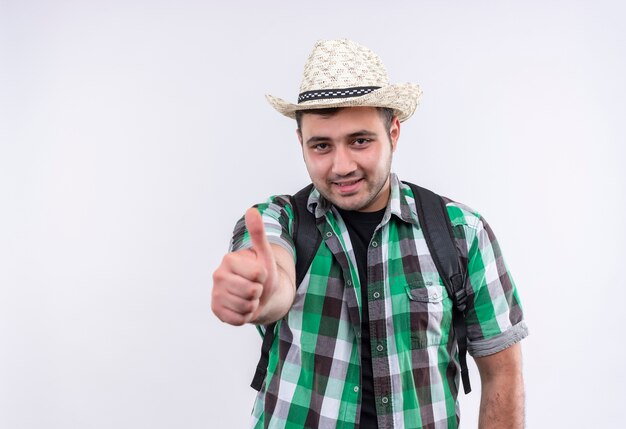 Young traveler man in checked shirt and summer hat with backpack smiling with happy face showing thumbs up standing over white wall
