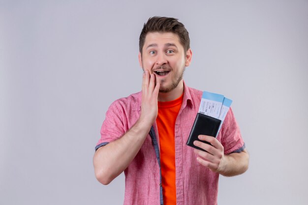 Young traveler man holding air tickets looking at camera amazed and surprised happy and exited standing over isolated white background