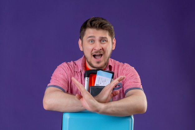 Free photo young traveler man holding suitcase and air tickets looking at camera amazed making stop gesture crossing hands standing over purple background