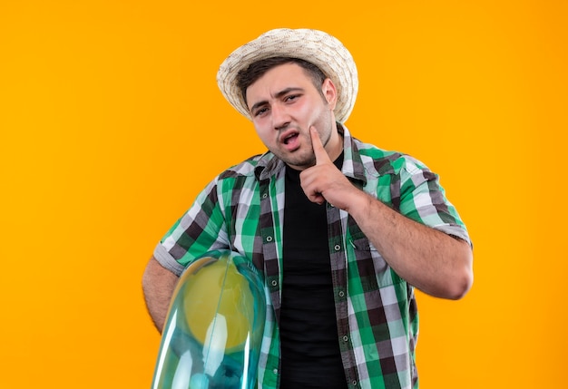 Молодой путешественник в клетчатой рубашке и летней шляпе держит надувное кольцо с задумчивым выражением лица над оранжевой стеной