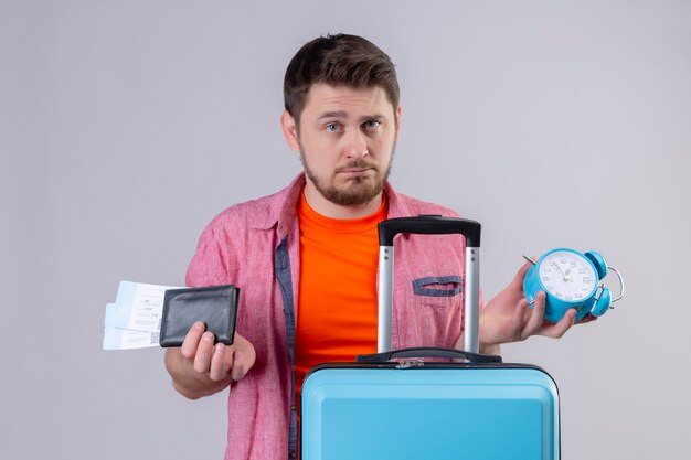 Young traveler man standing with suitcase holding air tickets and alarm clock looking at camera displeased standing over white background