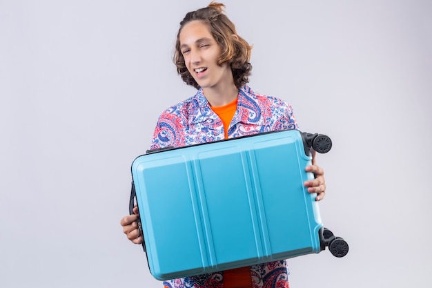 Young traveler man with blue suitcase looking confident smiling with happy face ready to travel standing over white background