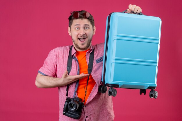 Young traveler man with camera holding suitcase looking at camera happy and exited smiling cheerfully standing over pink background