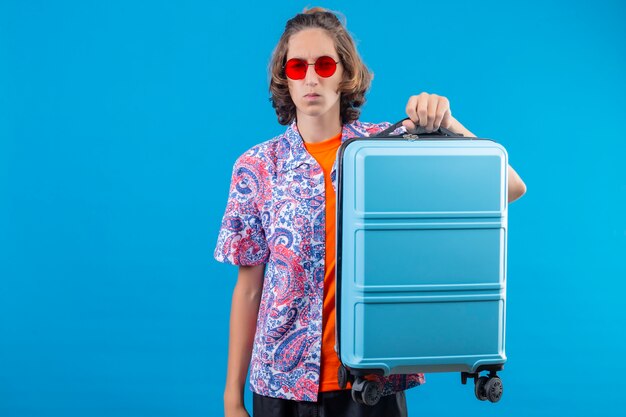 Молодой путешественник с чемоданом, держащий авиабилеты, выглядит смущенным, стоя на синем фоне