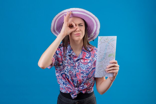 Young traveler woman in summer hat holding map doing ok sign looking through this sign with suspicious expression standing over blue background