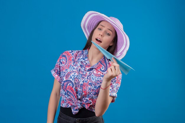 Молодой путешественник женщина в летней шляпе держит бумажный самолетик игривое и счастливое положение на синем фоне