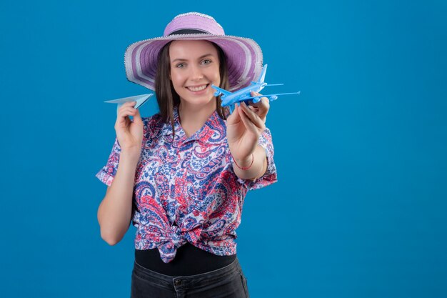 Young traveler woman in summer hat holding paper and toy airplanes, playing with them positive and happy over blue wall