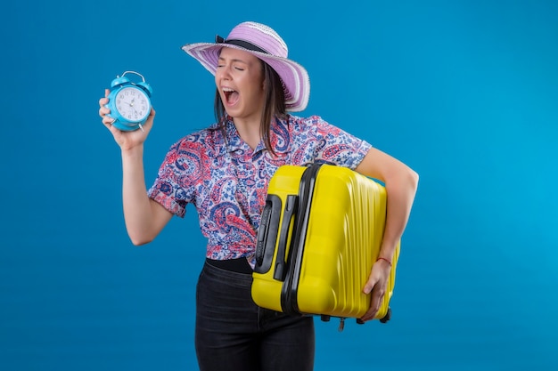 Young traveler woman in summer hat standing with yellow suitcase holding alarm clock annoyed and frustrated shouting with anger over blue background