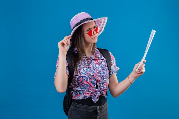 Молодая женщина-путешественница в летней шляпе в красных солнцезащитных очках держит карту, глядя в сторону с задумчивым выражением лица, думая, имея сомнения, стоя на синем фоне