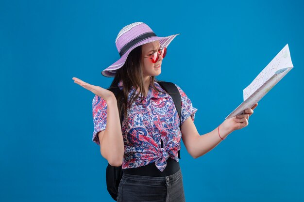 그것을보고지도를 들고 빨간 선글라스를 착용하는 여름 모자에 젊은 여행자 여자 파란색 배경 위에 서있는 아무 생각 개념도 오픈 팔과 혼동