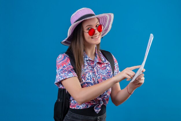 Молодая женщина-путешественница в летней шляпе в красных солнцезащитных очках держит карту, указывая пальцем на нее, весело улыбаясь, стоя на синем фоне