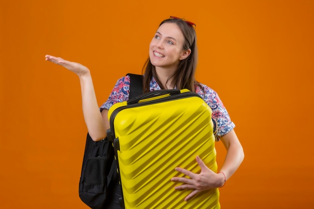 Молодая женщина-путешественница в красных солнцезащитных очках на голове, стоящая с рюкзаком, держащая чемодан, глядя в сторону и представляющая рукой что-то улыбающееся со счастливым лицом на оранжевом фоне