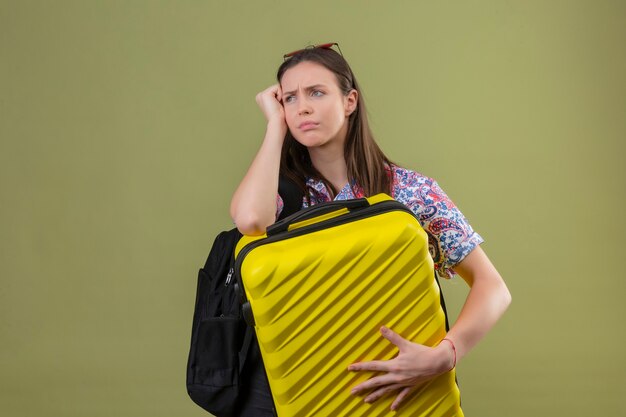 Young traveler woman wearing red sunglasses on head standing with backpack holding suitcase looking tired waiting with hand near face unhappy over green background