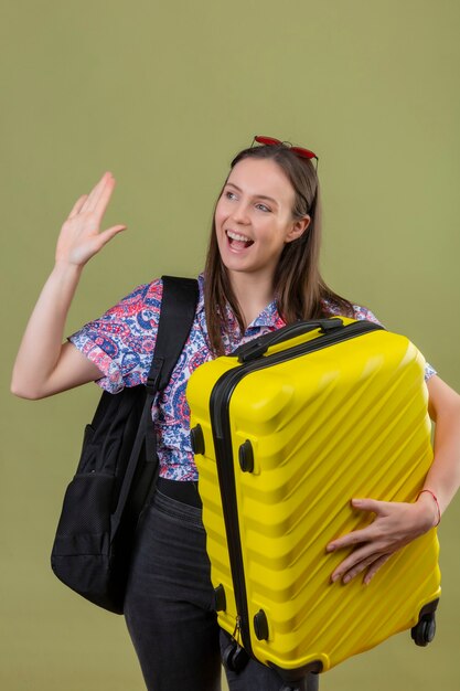 Young traveler woman wearing red sunglasses on head standing with backpack holding suitcase waving her hand while greeting or making goodbye gesture smiling with happy face over isolated green