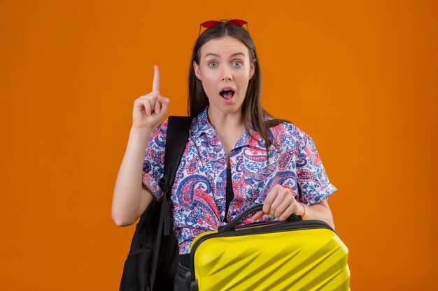 Молодая женщина путешественника нося красные солнечные очки на голове при рюкзак держа чемодан смотря удивленный и изумленный стоять с пальцем вверх, новая концепция идеи над оранжевой стеной