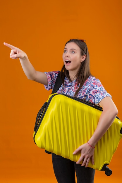 Young traveler woman wearing red sunglasses on head with backpack holding suitcase pointing with finger to the side looking surprised over orange wall