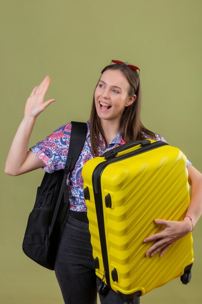 スーツケースを持ってバックパックで挨拶または別れのジェスチャーをしながら別れのジェスチャーをしながら孤立したグリーンを笑顔でバックパックを頭に赤いサングラスをかけている若い旅行者女性