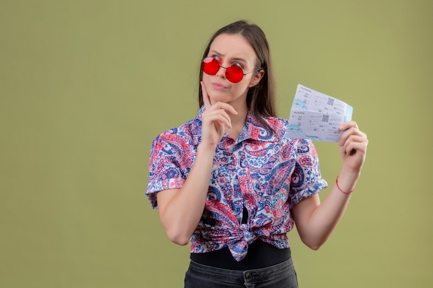 Young traveler woman wearing red sunglasses holding tickets looking aside with pensive expression displeased over green wall