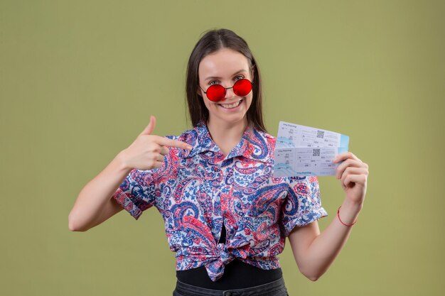 Молодая женщина путешественника нося красные солнечные очки держа билеты указывая палец к ним усмехаясь с счастливым лицом над зеленой стеной