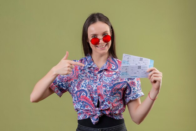 Young traveler woman wearing red sunglasses holding tickets pointing with finger to them smiling with happy face standing over green background