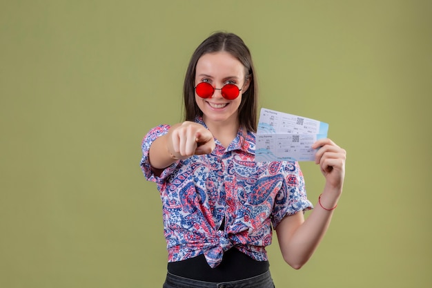 Young traveler woman wearing red sunglasses holding tickets smiling with happy face pointing with finger to camera standing over green background