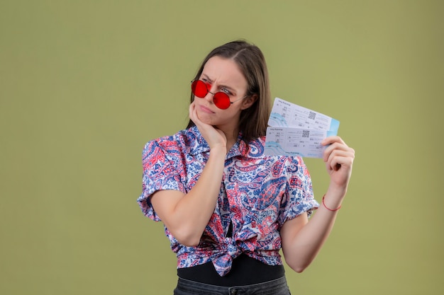 Young traveler woman wearing red sunglasses holding tickets with hand on chin displeased and thinking with pensive expression over green wall