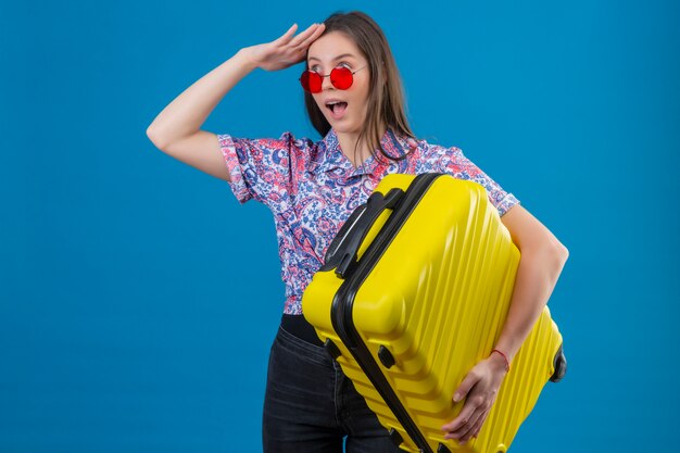 Молодая женщина путешественника нося красные солнечные очки держа желтый чемодан смотря далеко с рукой для того чтобы посмотреть что-то, положительный и удивленный над голубой стеной