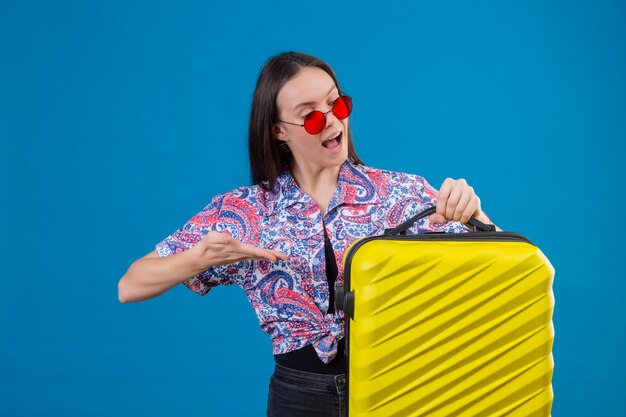 Int 파란색 벽에 즐거운 찾고 손 팔으로 가리키는 노란색 가방을 들고 빨간 선글라스를 착용하는 젊은 여행자 여자