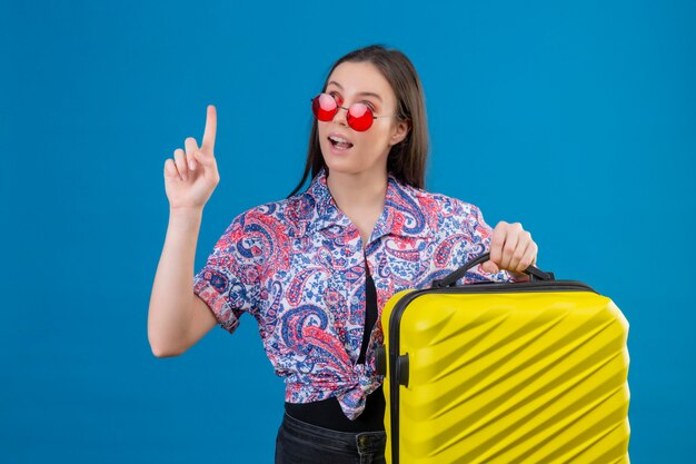 Young traveler woman wearing red sunglasses holding yellow suitcase with finger up looking confident having great idea over blue wall