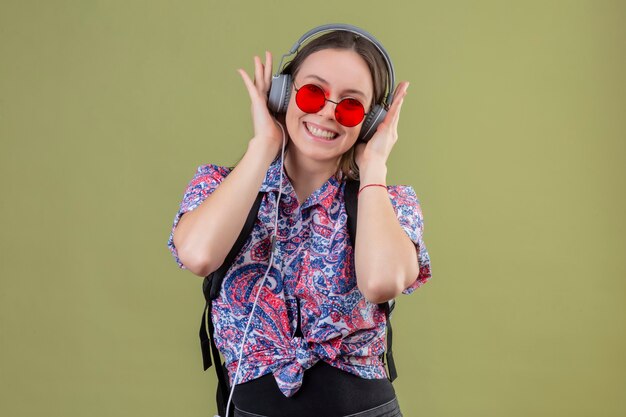 빨간 선글라스를 착용하고 배낭이 녹색 배경 위에 서있는 행복한 얼굴로 웃는 헤드폰을 사용하여 음악을 듣고 젊은 여행자 여자