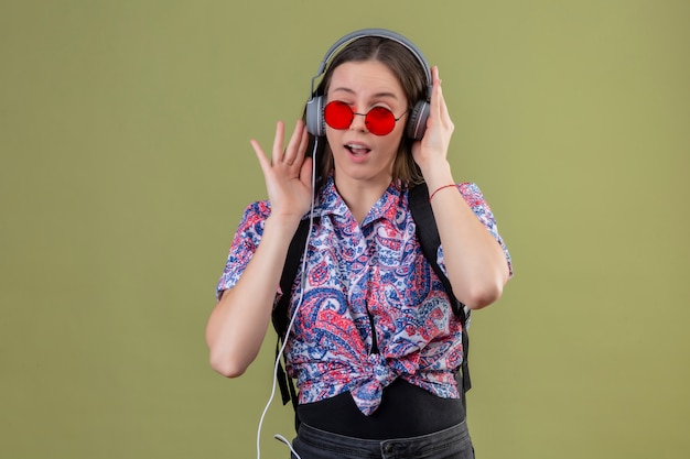 빨간 선글라스를 착용하고 배낭 녹색 벽을 통해 행복한 얼굴로 헤드폰을 사용하여 음악을 듣고 젊은 여행자 여자