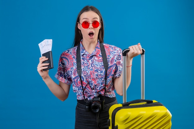 여권과 티켓을 들고 노란색 가방에 빨간 선글라스를 착용하는 젊은 여행자 여자는 파란색 벽을 놀라게하고 놀라게 찾고