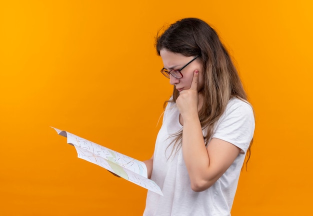 Young traveler woman in white t-shirt  holding map looking at it with pensive expression on face thinking standing over orange wall