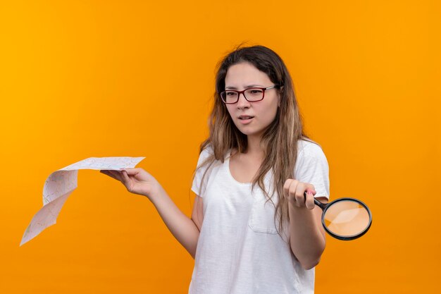 Young traveler woman in white t-shirt  holding map and magnifying glass looking, confused shrugging shoulders standing over orange wall