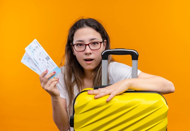 オレンジ色の壁の上に立って驚いて見えるスーツケースと航空券を保持している白いTシャツの若い旅行者の女性