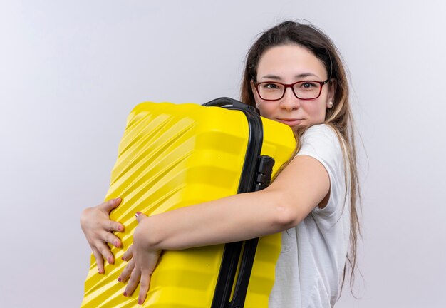 Young traveler woman in white t-shirt holding suitcase hugging her suitcase smiling positive and happy standing over white wall